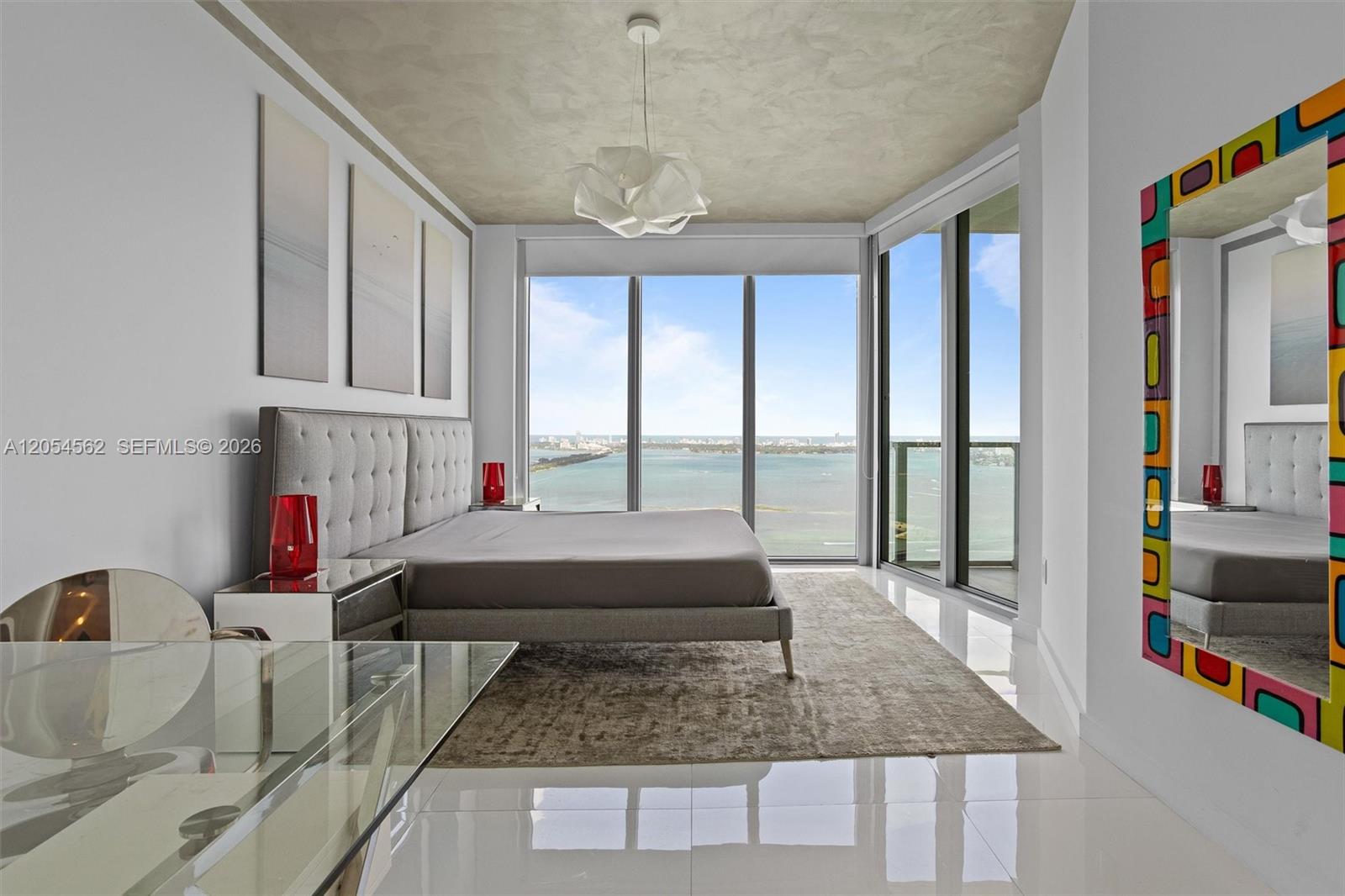 A modern bedroom features a gray tufted bed, floor-to-ceiling windows, and a colorful framed mirror — 2900 NE 7th Ave # 4205, Miami FL 33137, MIAMI, FL