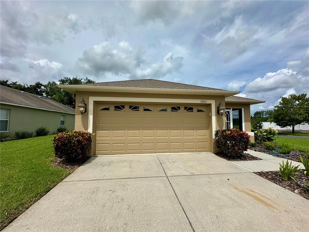 A tan single-story house features a two-car garage, a concrete driveway, and landscaped front bushes — 18861 WATER LILY LANE, HUDSON, FL