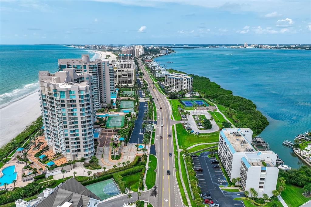 An aerial view shows high-rise condominiums, a road, tennis courts, and a beach along water — 1581 GULF BOULEVARD UNIT 203N, CLEARWATER BEACH, FL