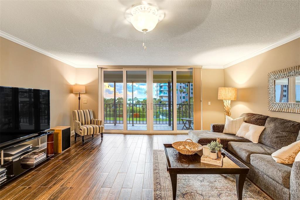 A living room features wood-look tile flooring, a sectional sofa, and sliding glass doors to a balcony — 1581 GULF BOULEVARD UNIT 203N, CLEARWATER BEACH, FL