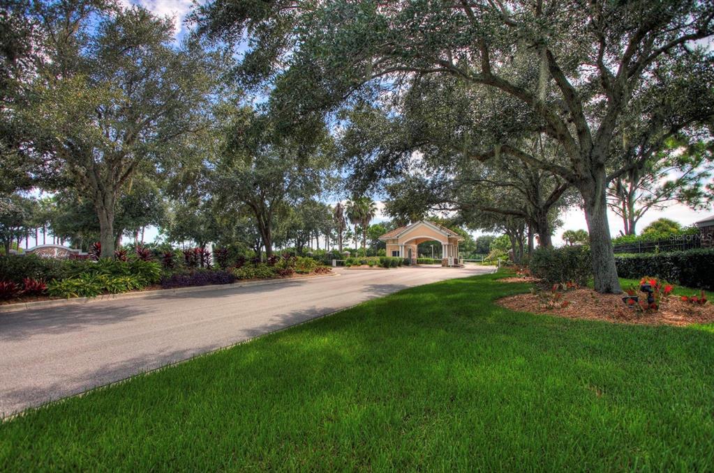 A paved road leads toward a gated community entrance under a canopy of large trees — 5301 MAHOGANY RUN AVENUE UNIT 1023, SARASOTA, FL
