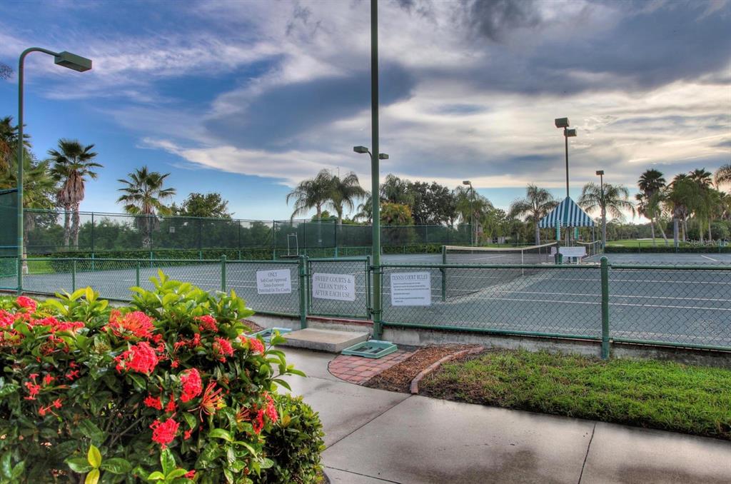 A fenced tennis court with a striped awning and surrounding palm trees under a cloudy sky — 5301 MAHOGANY RUN AVENUE UNIT 1023, SARASOTA, FL