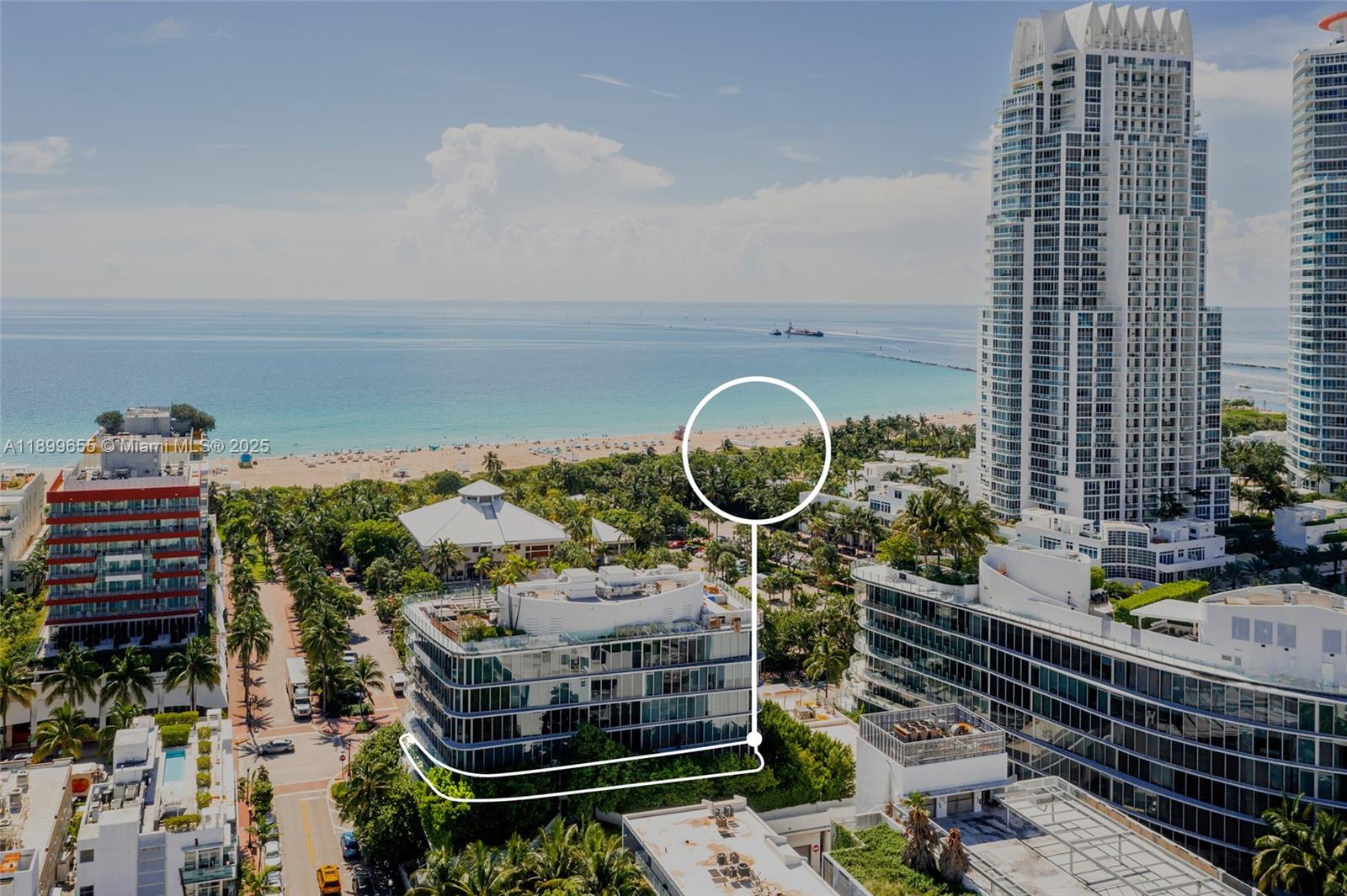 An aerial view shows a modern glass-walled building surrounded by lush trees near the beach — 1 Collins Ave # 207, Miami Beach FL 33139, MIAMI BEACH, FL