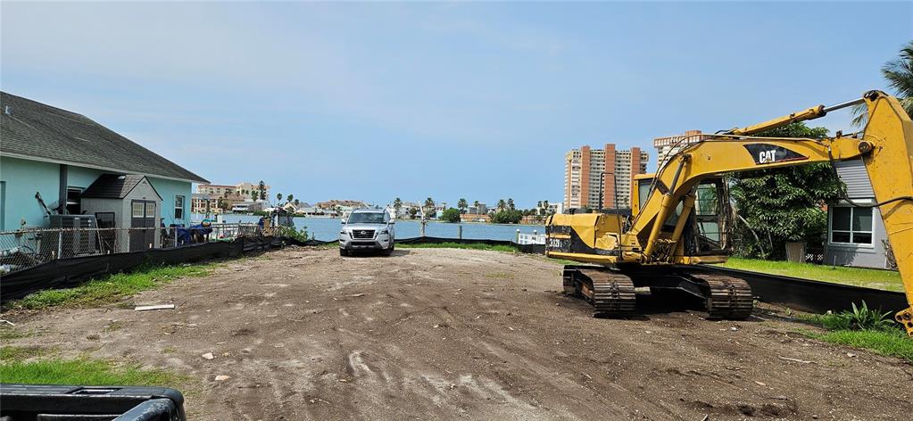 A yellow excavator sits on a dirt lot facing a waterfront view with buildings — 623 180TH AVENUE E, REDINGTON SHORES, FL