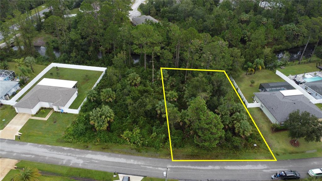 An aerial view shows a vacant, wooded residential lot outlined in yellow between two houses — 98 SLOGANEER TRAIL, PALM COAST, FL