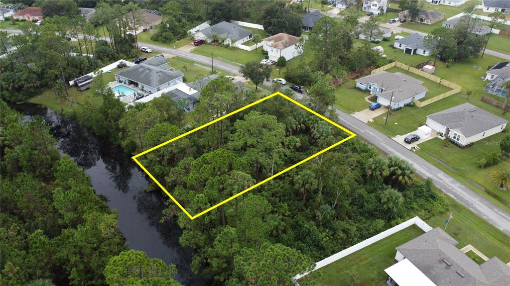 An aerial view shows a vacant, wooded residential lot outlined in yellow next to a canal — 98 SLOGANEER TRAIL, PALM COAST, FL