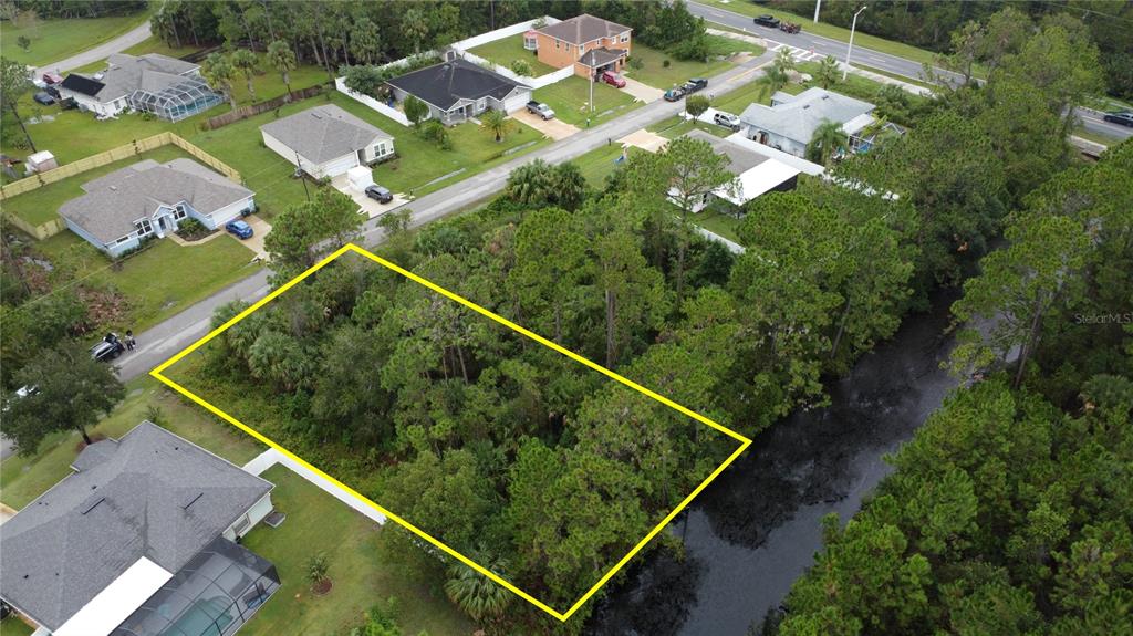 An aerial view shows a vacant, wooded residential lot outlined in yellow next to a canal — 98 SLOGANEER TRAIL, PALM COAST, FL