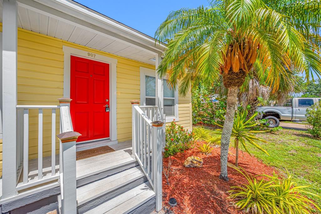 A yellow house exterior features a bright red front door and a small wooden porch — 907 18TH AVENUE W, PALMETTO, FL