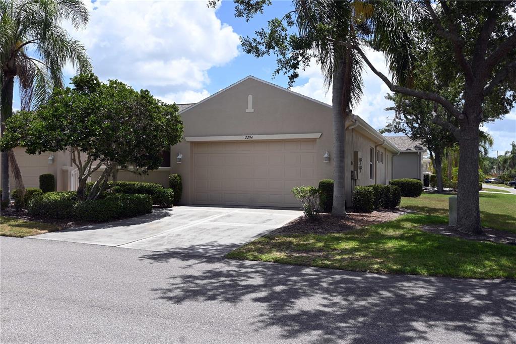 A beige house exterior features a two-car garage, a concrete driveway, and surrounding landscaping — 2294 SIFIELD GREENS WAY UNIT 2294, SUN CITY CENTER, FL