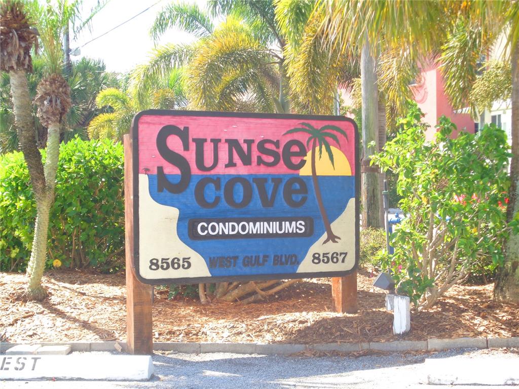 A wooden sign for Sunset Cove Condominiums stands outdoors surrounded by palm trees and bushes — 8565 W GULF BOULEVARD UNIT 11S, ST PETERSBURG, FL