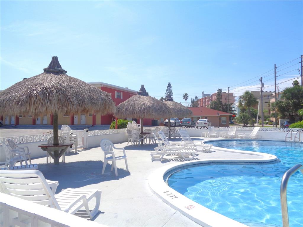 A swimming pool area features tiki umbrellas, white plastic lounge chairs, and a surrounding building — 8565 W GULF BOULEVARD UNIT 11S, ST PETERSBURG, FL