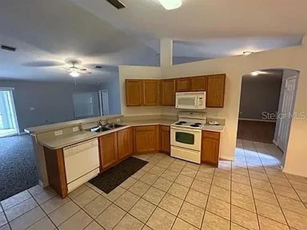 A kitchen with wood cabinets, white appliances, and tiled floors overlooks an adjacent living area — 2399 ORACLE LANE, NORTH PORT, FL
