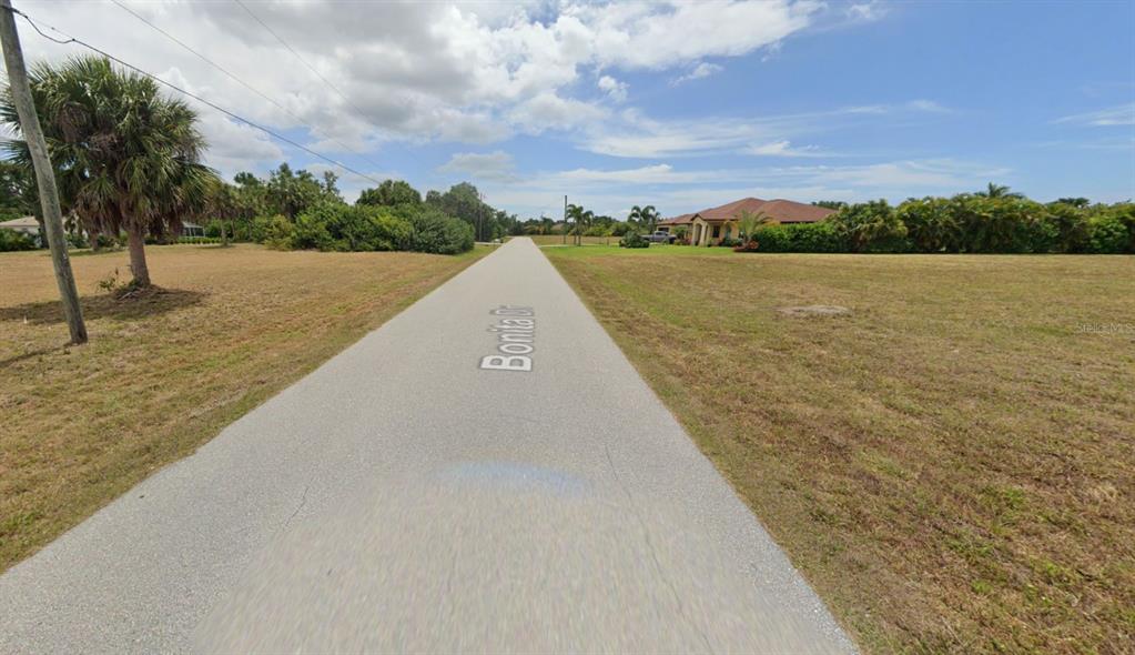 A paved road stretches through a grassy, undeveloped residential area under a cloudy sky — 103 BONITA DRIVE, ROTONDA WEST, FL