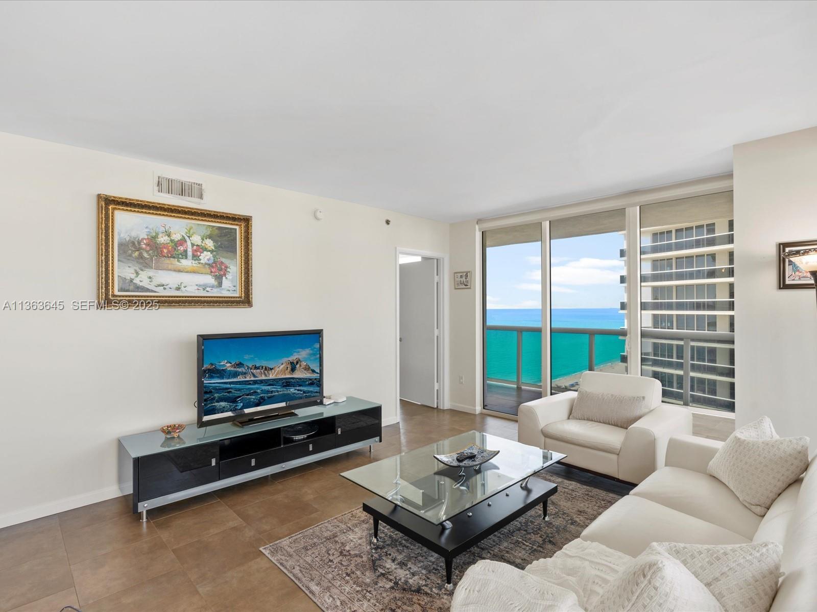 A living room features a television, white sofa, glass coffee table, and ocean views — 1830 S Ocean Dr, Hallandale Beach FL 33009, HALLANDALE BEACH, FL