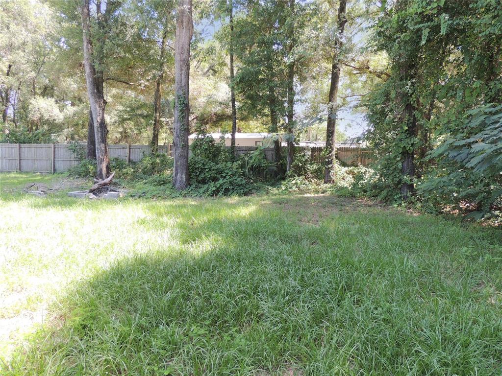 A grassy backyard features a wooden fence, several tall trees, and a small fire pit — 7439 SE 78TH PLACE, TRENTON, FL
