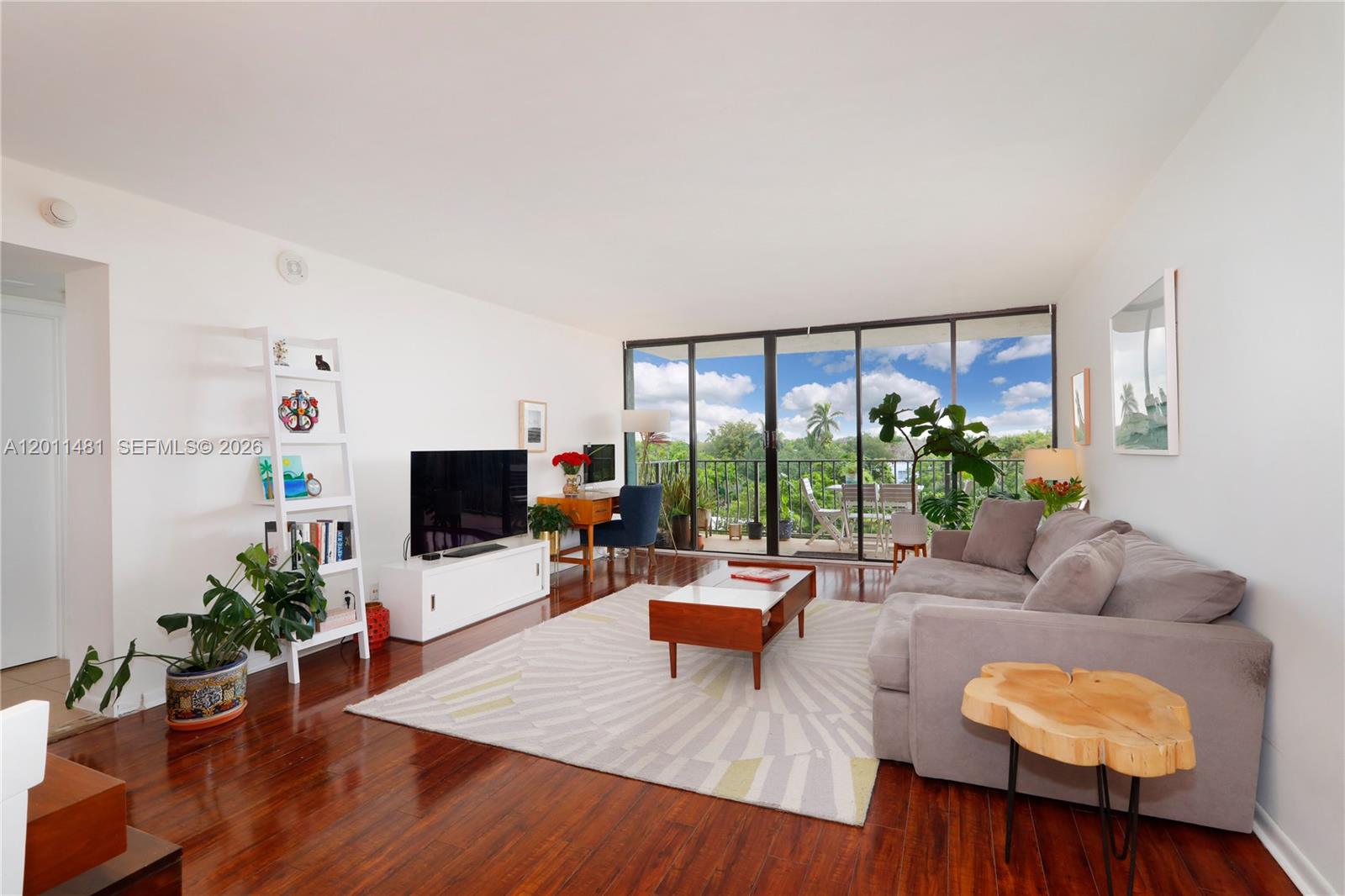 A living room with hardwood floors, a television, a desk, and a balcony with greenery — 780 NE 69th St # 404, Miami FL 33138, MIAMI, FL