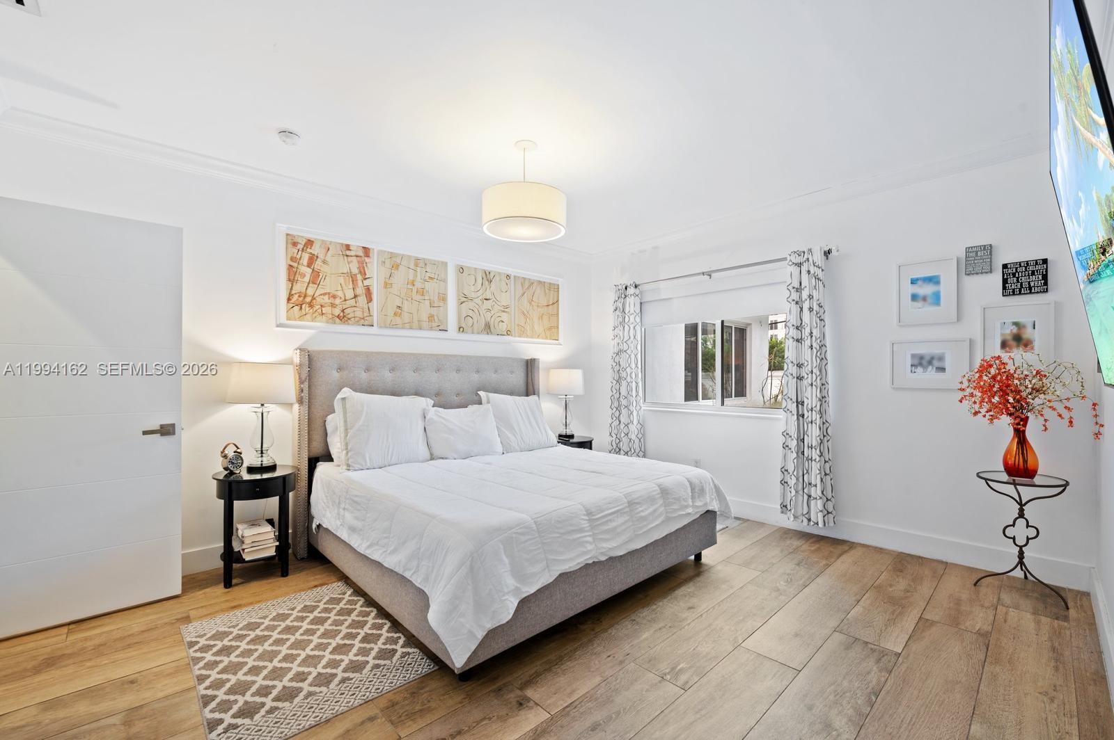 A bedroom features a tufted gray bed, wood-look flooring, and a window with patterned curtains — 3551 E Glencoe St, Miami FL 33133, MIAMI, FL