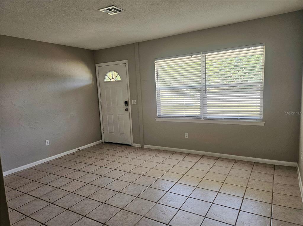 An empty room features beige tile flooring, gray walls, a white front door, and windows — 7000 BRENTFORD ROAD, SARASOTA, FL