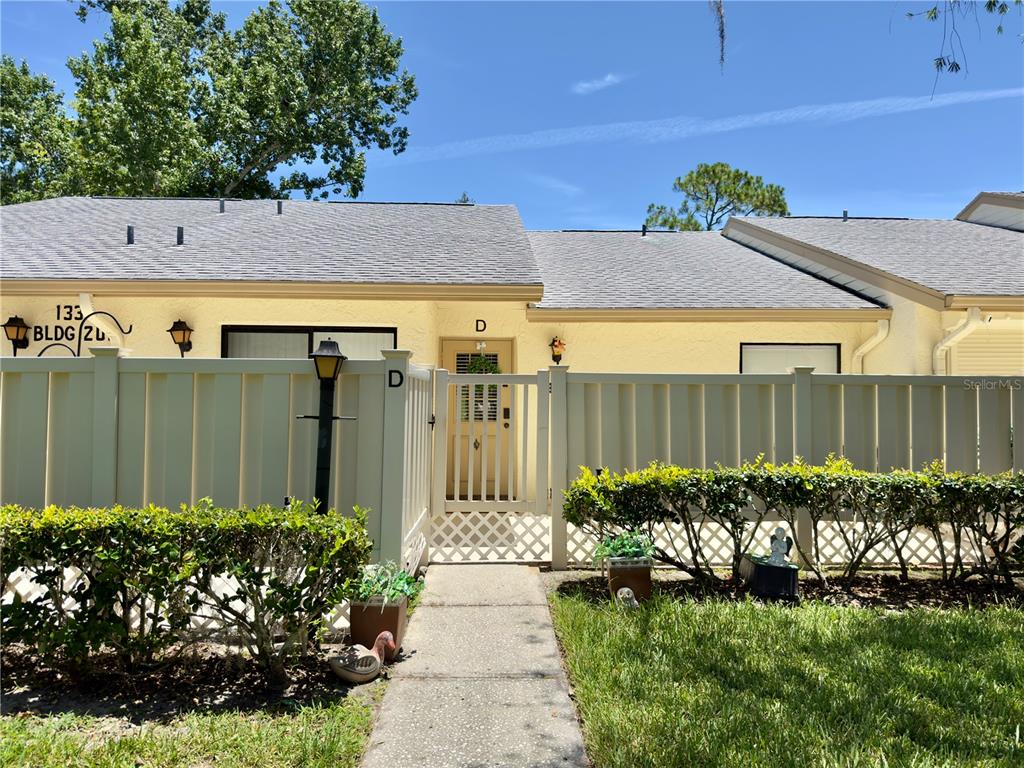 A beige, single-story residential building features a white privacy fence and a gated courtyard entrance — 1333 SHADY PINE WAY UNIT D, TARPON SPRINGS, FL