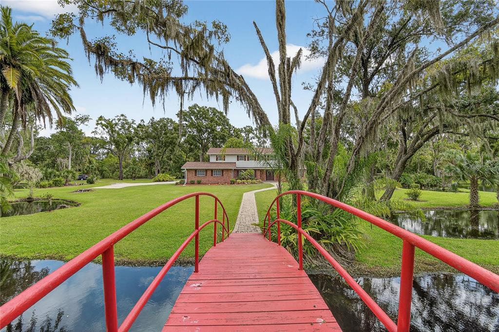 A red arched footbridge crosses a pond toward a two-story brick house on a lawn — 1502 AZALEA STREET, PLANT CITY, FL