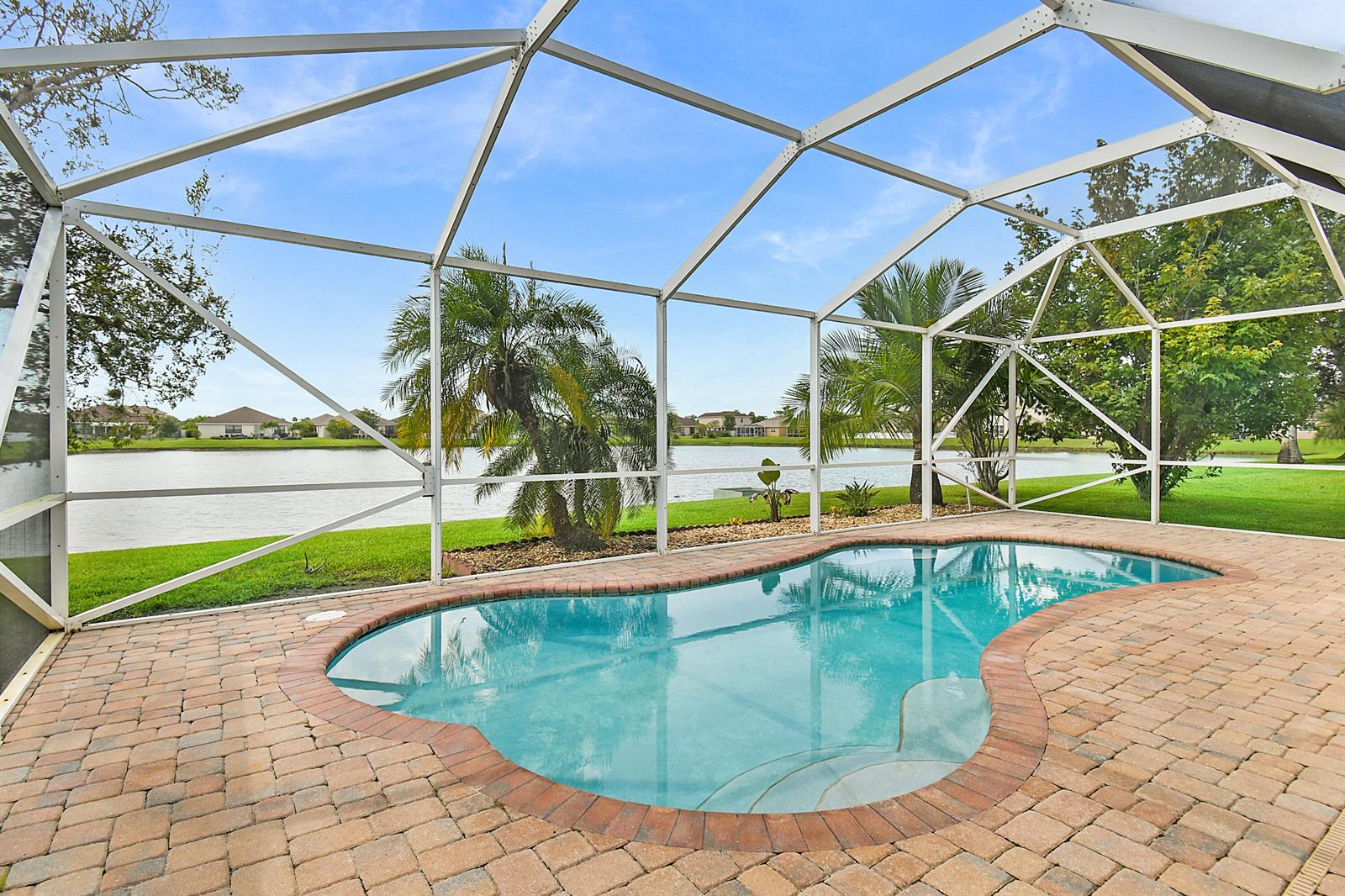 A swimming pool with a brick deck is enclosed by a screen cage overlooking a lake — 9902 E Verona Circle Circle, Vero Beach FL 32966, VERO BEACH, FL