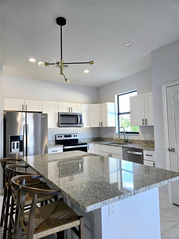 A kitchen features white cabinets, granite countertops, a stainless steel refrigerator, and a modern chandelier — 1819 NW 21ST STREET, CAPE CORAL, FL