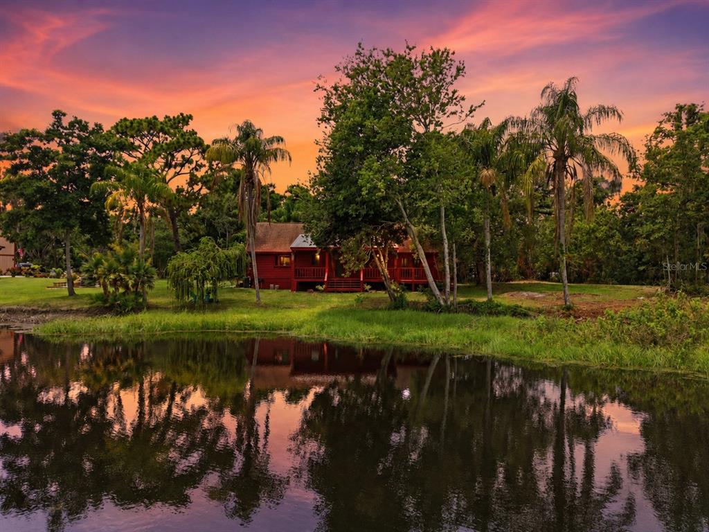 A red house sits behind a pond surrounded by trees under a vibrant sunset sky — 6409 CHISWICK COURT, NEW PORT RICHEY, FL
