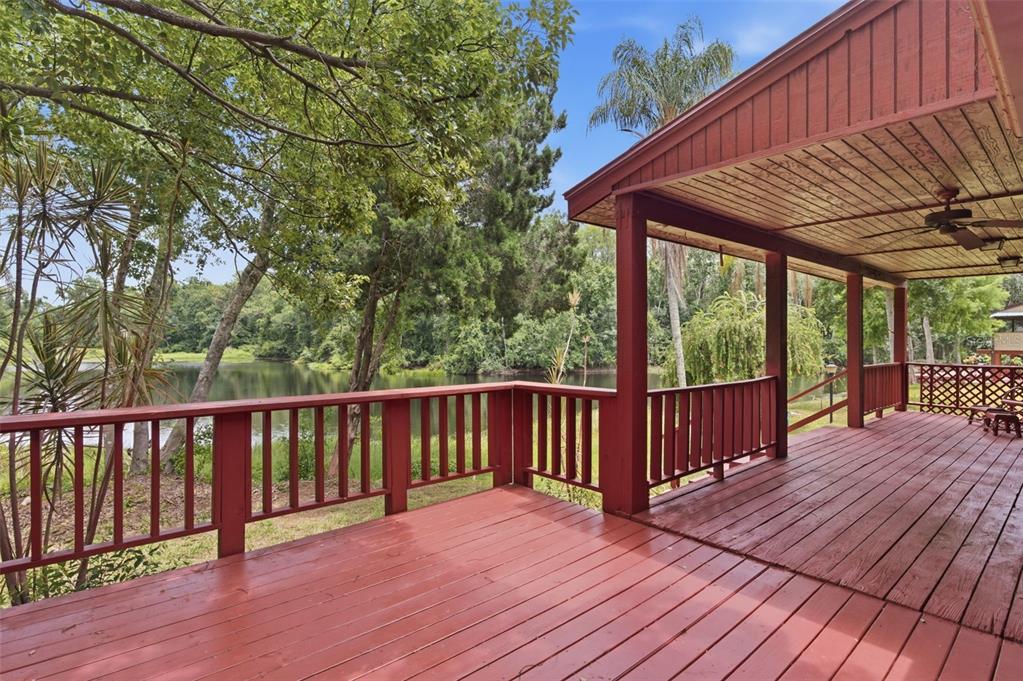 A red wooden deck with a covered porch overlooks a lake surrounded by trees — 6409 CHISWICK COURT, NEW PORT RICHEY, FL
