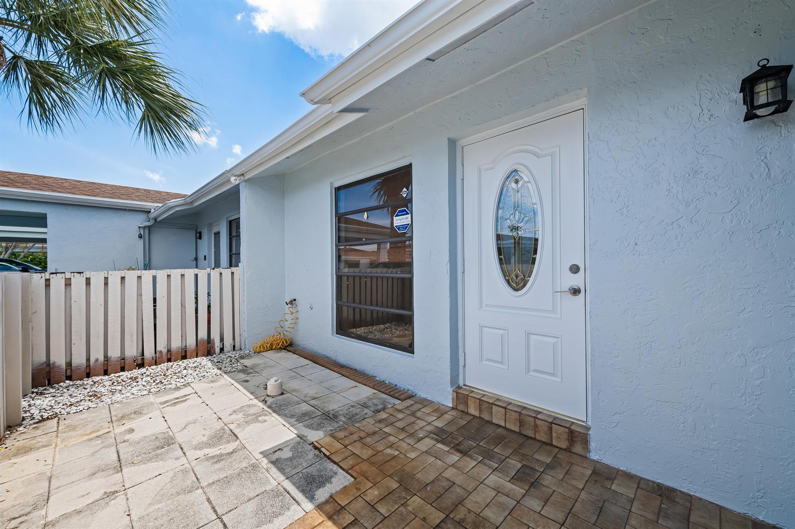 A light blue house exterior features a white front door, a window, and paved patio — 6673 Moonlit Drive, Delray Beach FL 33446, DELRAY BEACH, FL