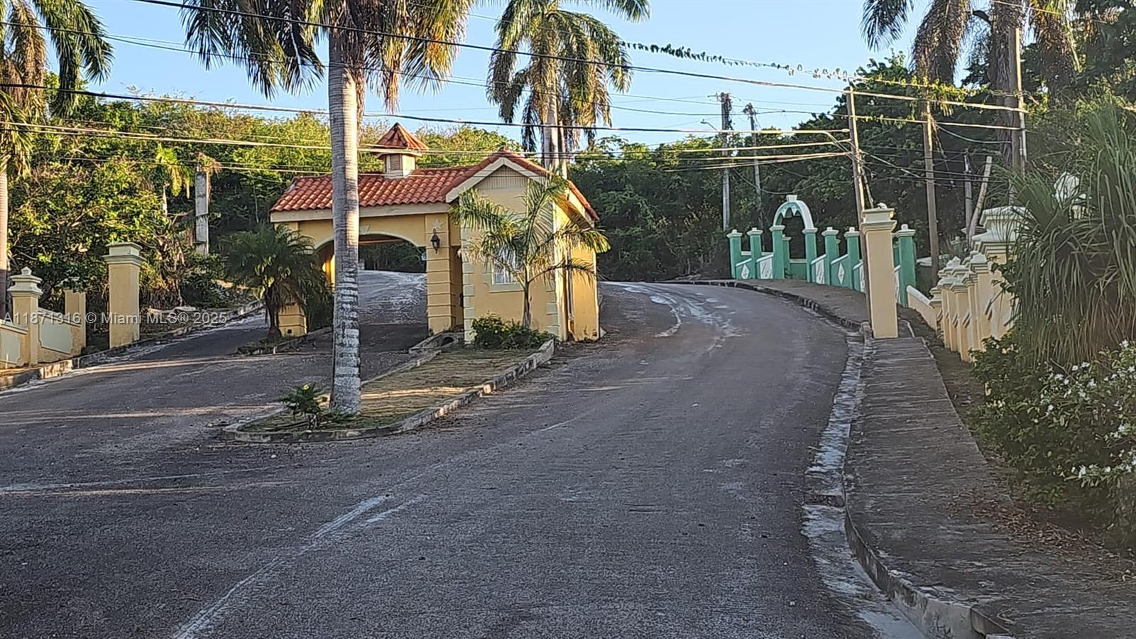 A paved road leads toward a gated entrance with a yellow guardhouse and decorative fencing — 00 Heavenly Heights Hanover,Jamaica, NA, NA