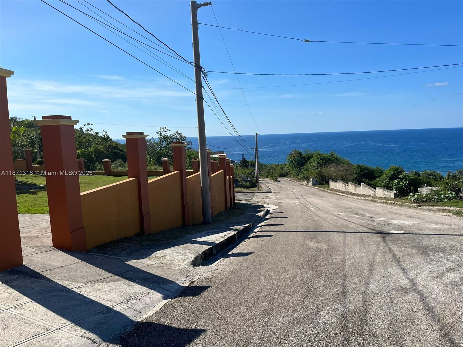 A paved road slopes downward toward the ocean beside a long, orange-walled property boundary — 00 Heavenly Heights Hanover,Jamaica, NA, NA