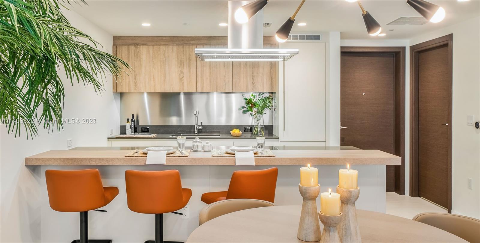 A modern kitchen features light wood cabinets, a stainless steel backsplash, and an island with orange stools — 1000 Brickell Plaza # 2711, Miami FL 33131, MIAMI, FL