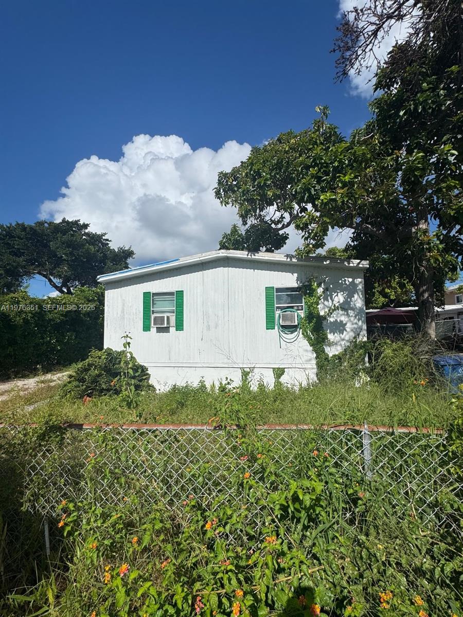 A white mobile home with green shutters sits behind a chain-link fence and overgrown vegetation — 13 Judy Pl, Key Largo FL 33037, KEY LARGO, FL