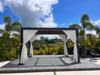 A dark-framed pergola with white curtains stands on a stone patio overlooking lush greenery — Lot 8 PR-159 Km 1.9 Inter BO. UNIBON, MOROVIS, PR
