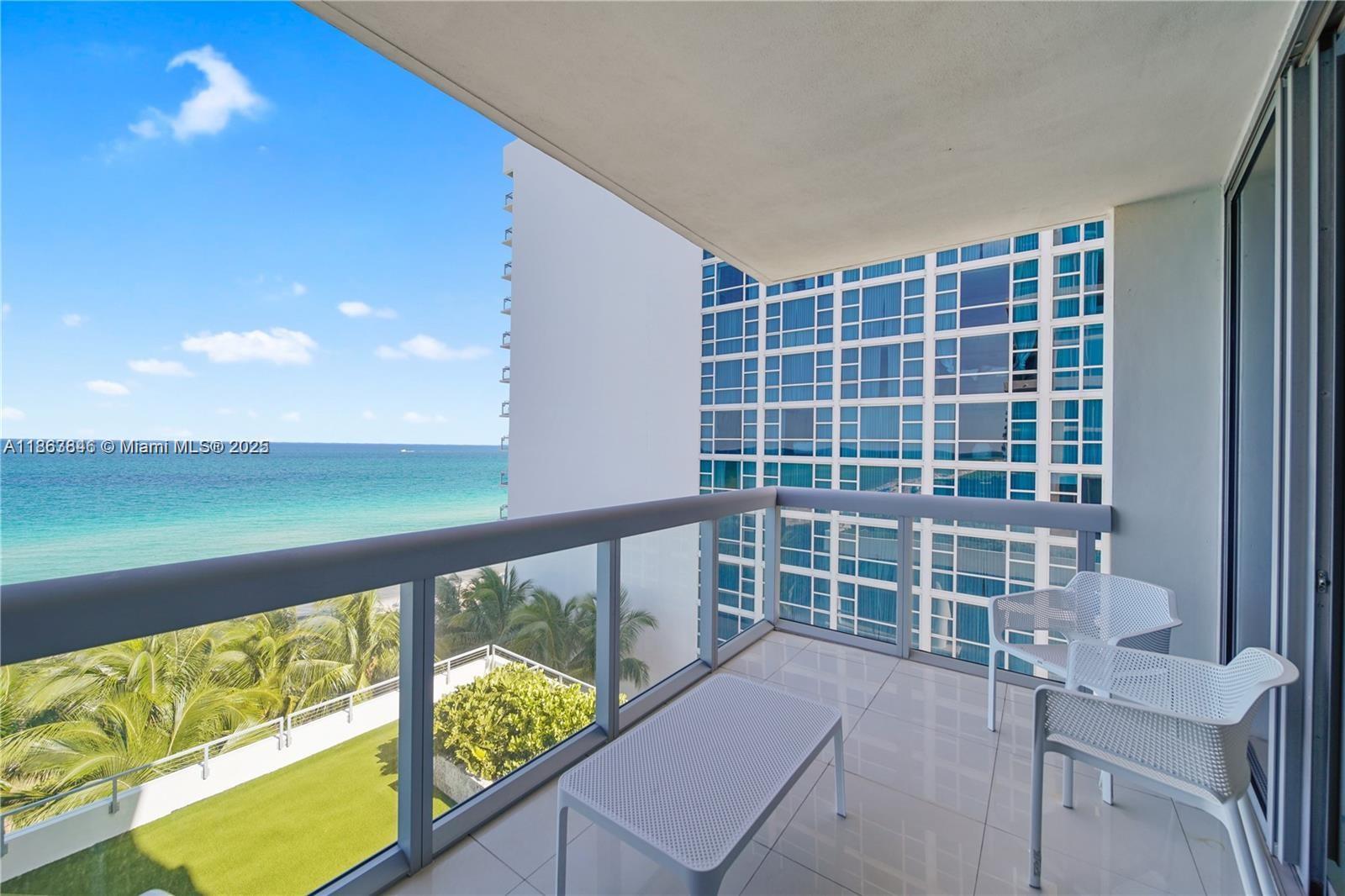 A balcony with white furniture overlooks the ocean and a neighboring high-rise building — 6899 Collins Ave # 808, Miami Beach FL 33141, MIAMI BEACH, FL