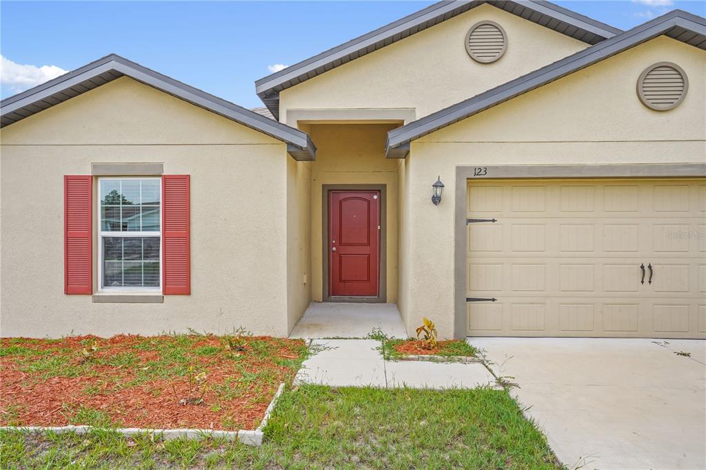 A single-story house with a beige stucco exterior, a red front door, and a garage — 123 VIOLET COURT, POINCIANA, FL