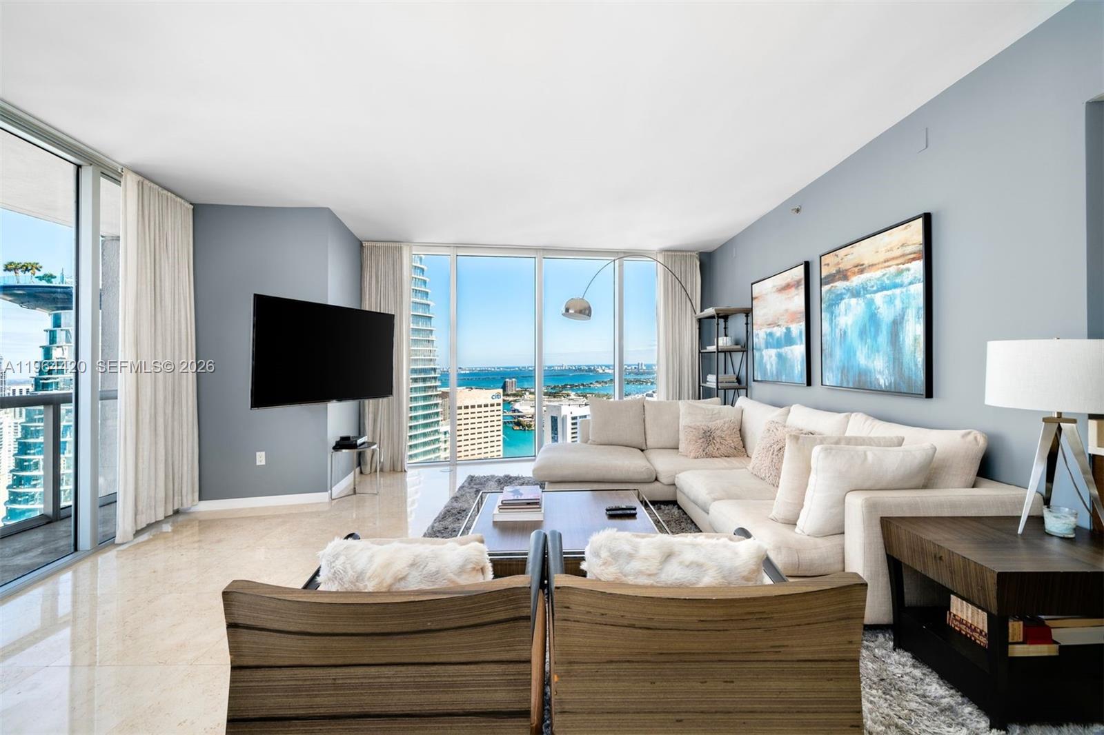 A modern living room features a sectional sofa, two wooden chairs, and floor-to-ceiling windows — 465 Brickell Ave # 5302, Miami FL 33131, MIAMI, FL