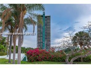 A tall residential high-rise building stands behind palm trees and flowering bushes under a cloudy sky — 780 NE 69th St # 1406, Miami FL 33138, MIAMI, FL