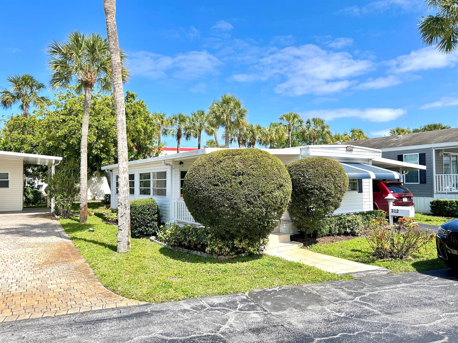 A white mobile home with a paved driveway, manicured shrubs, and palm trees under blue skies — 312 Hibiscus Drive, Deerfield Beach FL 33442, DEERFIELD BEACH, FL