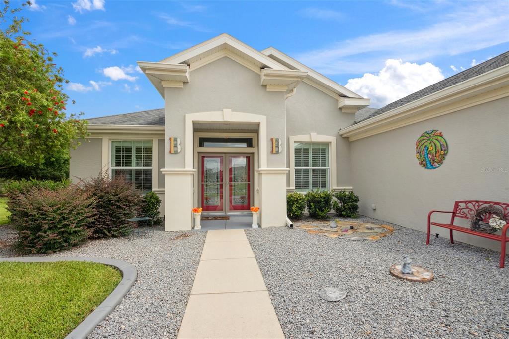 A single-story house exterior features a gray stucco facade, red double doors, and gravel landscaping — 13944 SE 96TH CIRCLE, SUMMERFIELD, FL