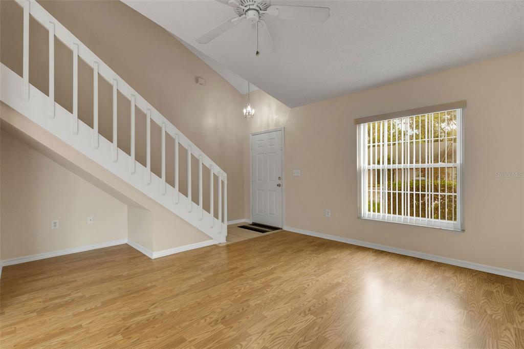 An empty living room features light wood-look flooring, a white staircase, and a large window — 1084 MEADOWOOD POINTE ROAD, LAKELAND, FL