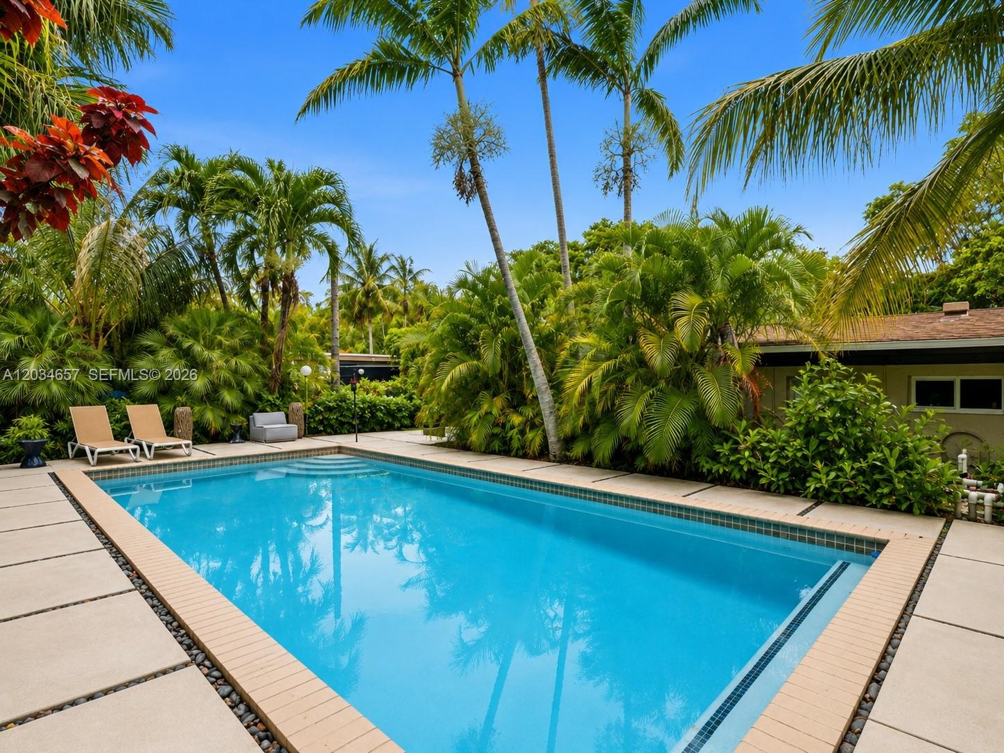 A rectangular swimming pool is surrounded by a paved patio and lush tropical landscaping — 1850 Keystone Blvd, North Miami FL 33181, NORTH MIAMI, FL