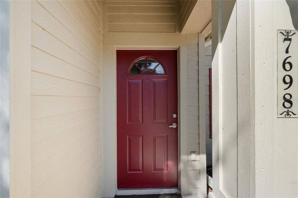 A red front door is set within a beige exterior wall with vertical house numbers — 7698 PLANTATION CIRCLE, BRADENTON, FL