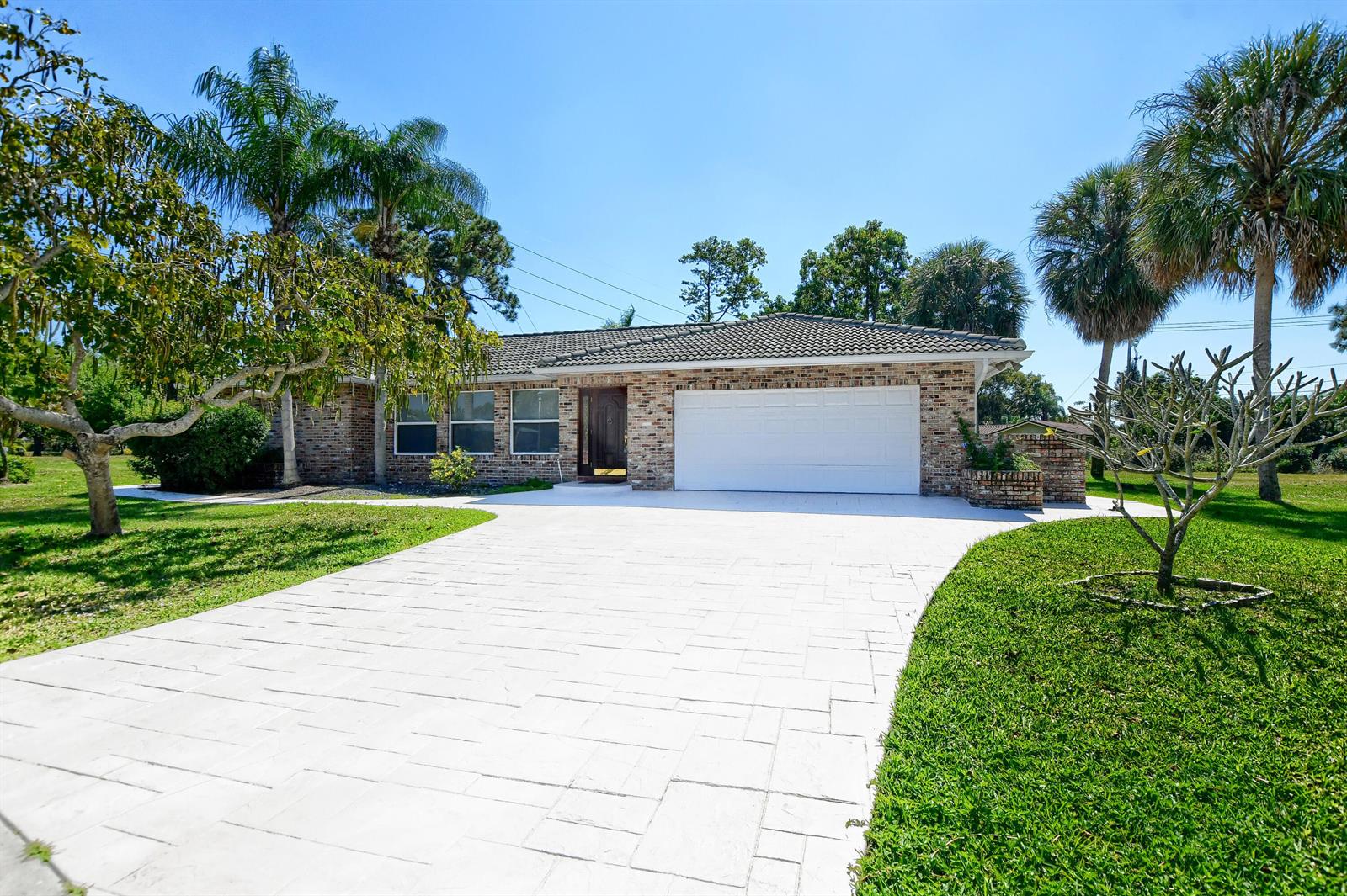A single-story brick house features a white garage door, a paved driveway, and palm trees — 2334 Timbercreek Circle NW, Boca Raton FL 33431, BOCA RATON, FL