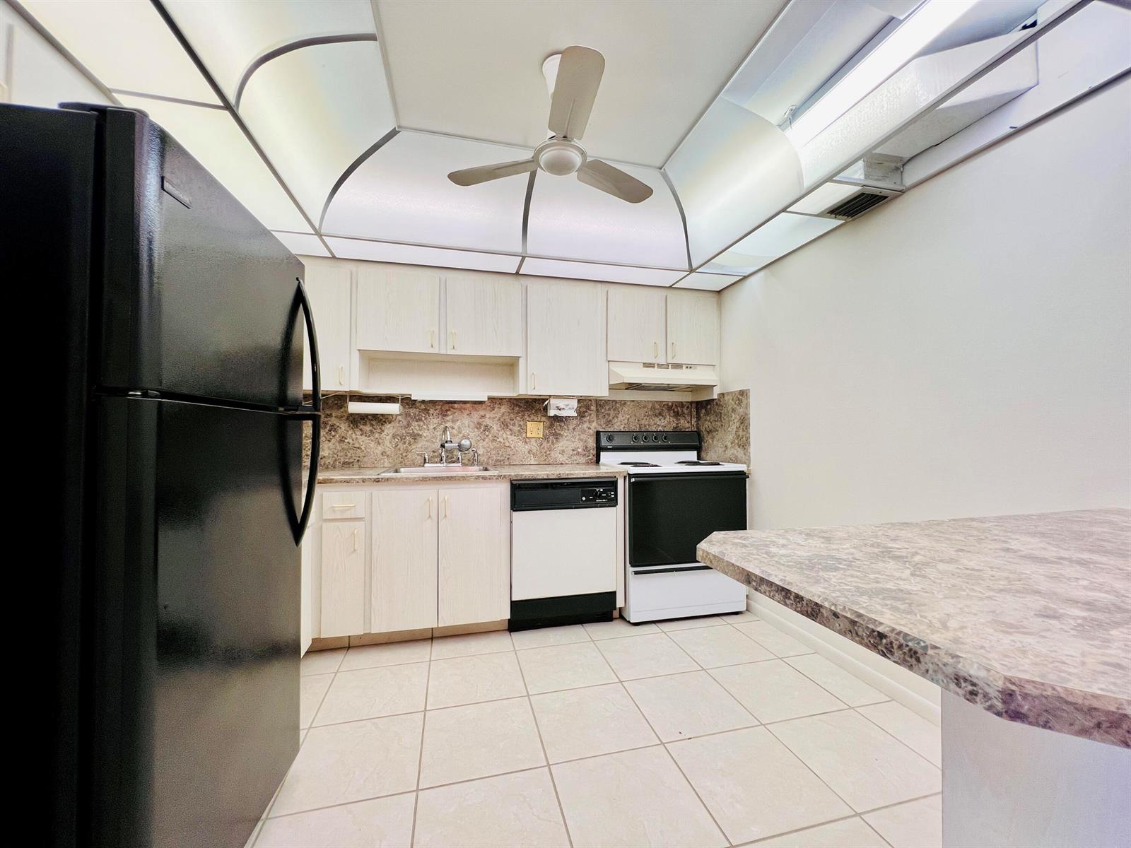 A kitchen features light wood cabinets, a black refrigerator, a white stove, and tile flooring — 107 Wellington D, West Palm Beach FL 33417, WEST PALM BEACH, FL
