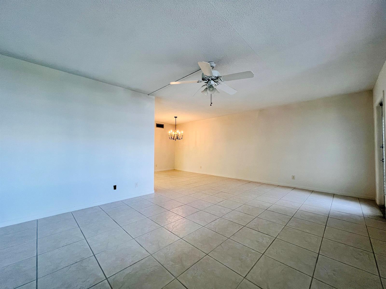 An empty room with light-colored tile flooring, a ceiling fan, and a distant chandelier — 107 Wellington D, West Palm Beach FL 33417, WEST PALM BEACH, FL