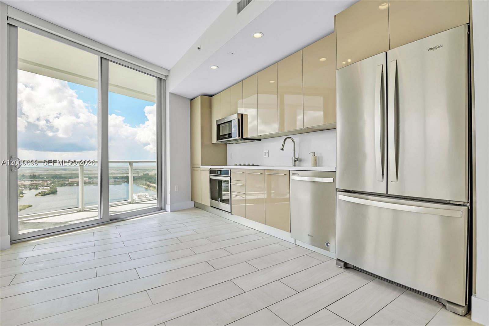 A modern kitchen features glossy beige cabinets, stainless steel appliances, and a balcony with water views — 2000 Metropica Way # 2204, Sunrise FL 33323, SUNRISE, FL