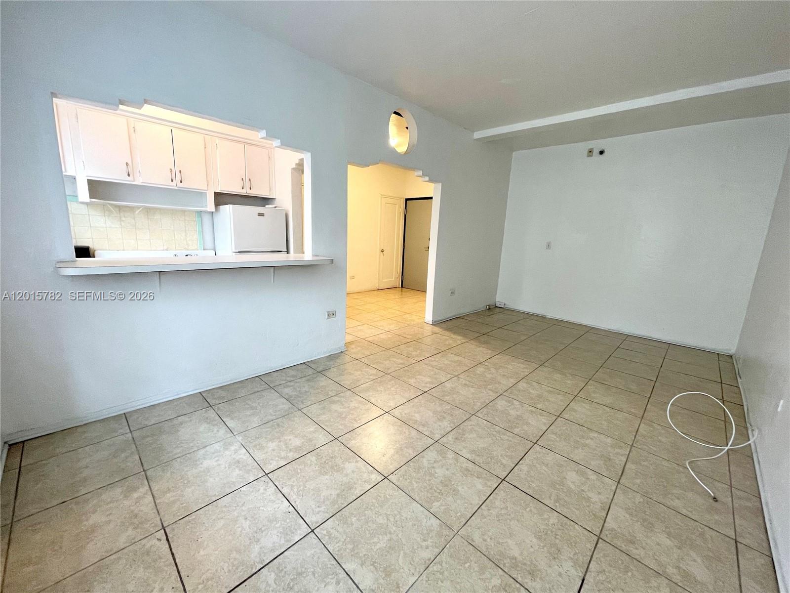 An open living area features beige tile flooring, a kitchen pass-through window, and white walls — 901 Collins Ave # 103, Miami Beach FL 33139, MIAMI BEACH, FL