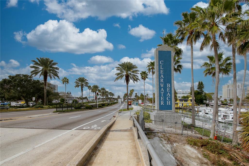 A Clearwater Beach sign stands beside a sidewalk, road, and marina under a blue sky — 1301 GULF BOULEVARD UNIT 201, CLEARWATER BEACH, FL