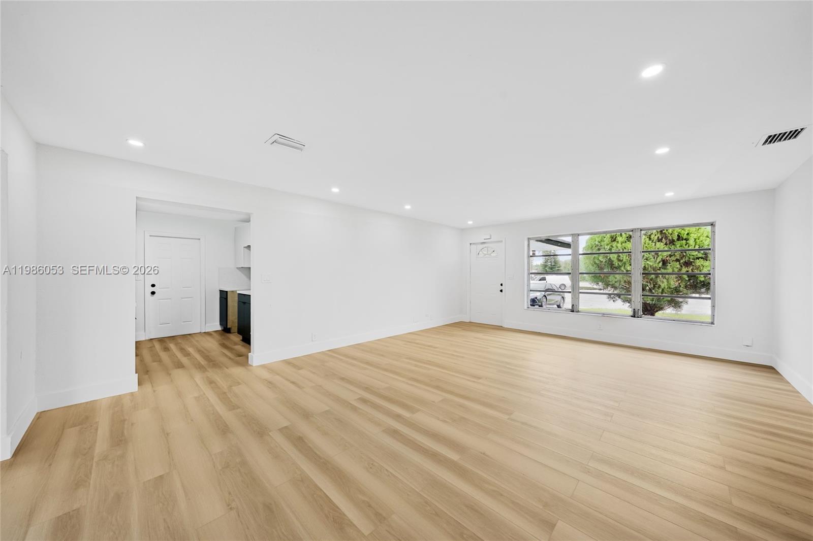 A bright, empty living room features light wood-look flooring, white walls, and a large window — 1353 NW 62nd Way, Margate FL 33063, MARGATE, FL